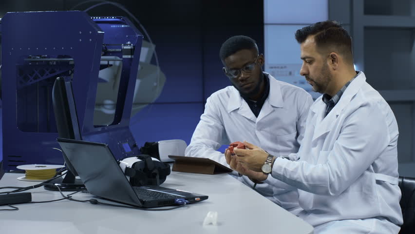 Multiethnic man in white gown discussing usage of three-dimensional printing in manufacturing process while exploring prototype. | Shutterstock HD Video #1010067332