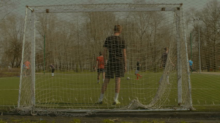 Young football forward dribbling, getting past defender and scoring a goal during football training match. View from behind goal. Fottball team playing soccer on sports field.