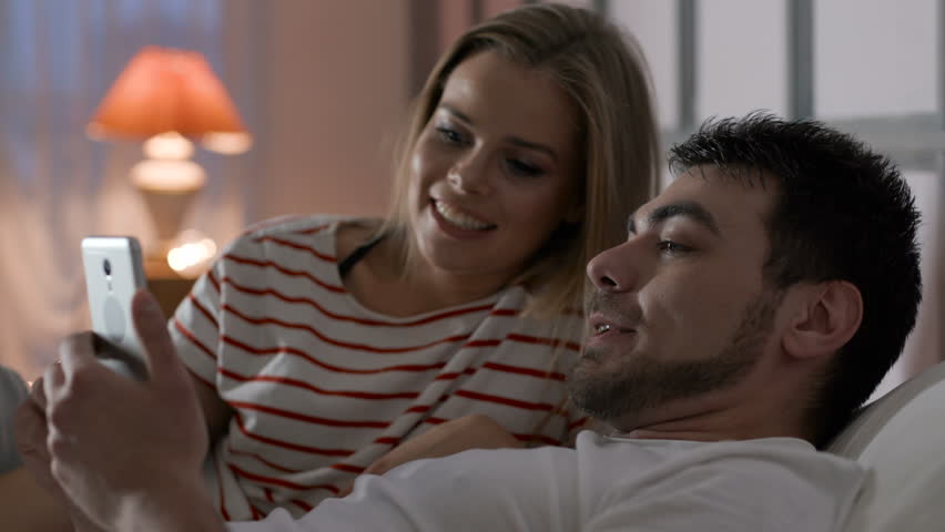 Caucasian modern newlyweds holding smart device in hand indoors. Relation of husband and wife or lovers. Romance of male person and cute female girl. Relaxation with digital social networking everyday