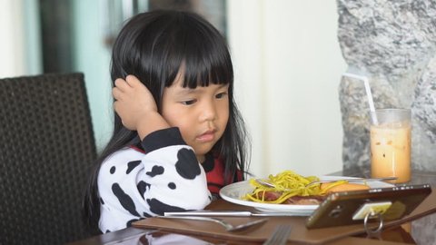 Asian children cute or kid girl smile with happy eating food with iced tea for breakfast or lunch and watch cartoon on smartphone because nomophobia or attention deficit hyperactivity disorder ADHD