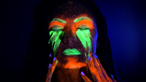 Portrait of girl smears and rubs fluorescent paint under eyes. Dye glowing near UV black light. Woman with braids in neon light. Night club, party, halloween psychedelic concepts.