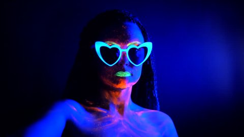 Fashion sexy dancer with heart shaped glasses in neon light. Fluorescent makeup glowing under ultraviolet light. Night club, party, psychedelic concepts. Mysterious woman with UV painting