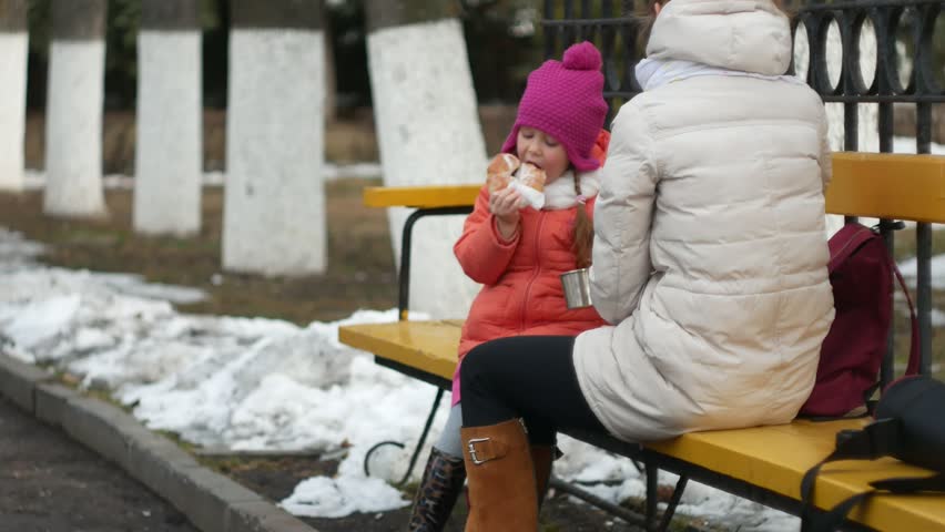 A beautiful girl and her mom are sitting on a park bench and eating a delicious dessert and drinking tea from a thermos in early spring 4k.