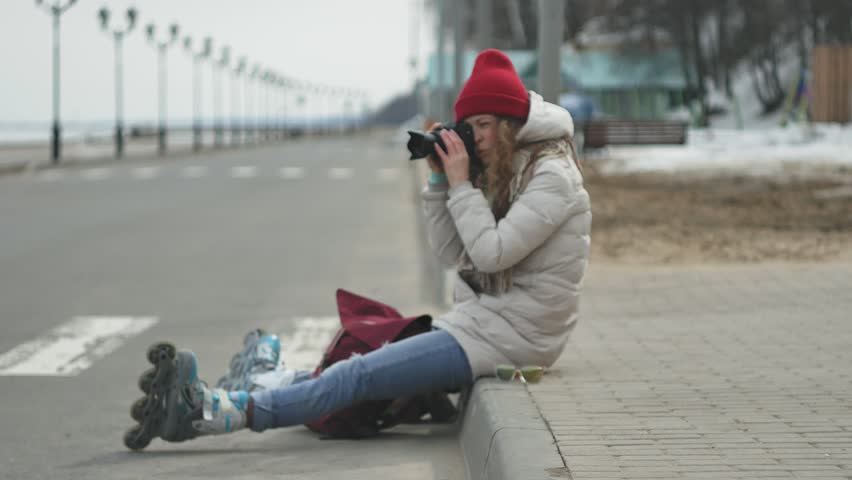Young beautiful woman in red hat wearing sporty warm clothes and rollers, sitting on the asphalt road and taking pictures on a vintage camera 4k
