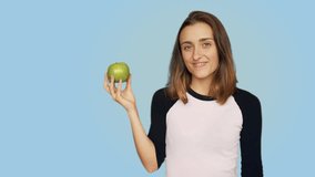 Cute and pretty beautiful teenager or young hipster woman, fitness or yoga practice,pastel blue background bites juicy ripe green apple, enjoys fresh fruit health benefits and smiles
