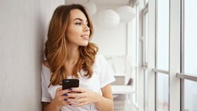 Happy brunette woman standing near the window while holding smartphone and looking at the camera at home