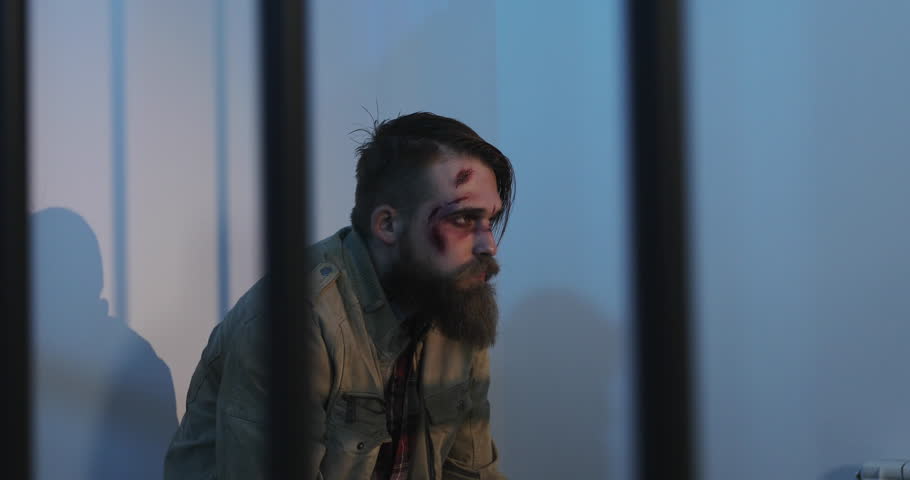 A wounded man with a beard sits behind a prison grate. A battered prisoner with wounds on his face.