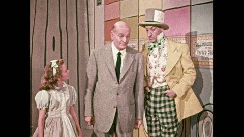 1950s: Girl walks down hallway with man and mad hatter. Woman with hat over head walks behind them, man talks, stops in front of large film reel. Mad hatter and hat covered woman operate projector.