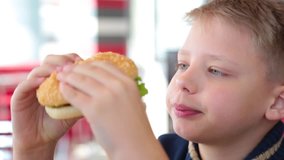 Closeup view of white child eating juicy big hamburger at fast food restaurant. Real time full hd video footage.