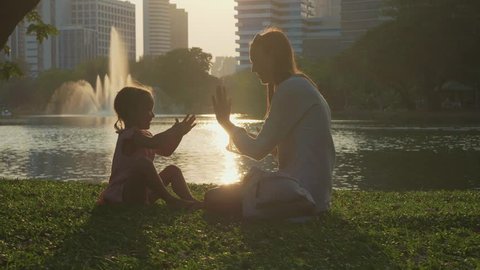 Mother having fun with her little cute daughter. Mom and child playing clapping game in the park with skyscrapers on the background at sunset and mother start tickling daughter in slow motion