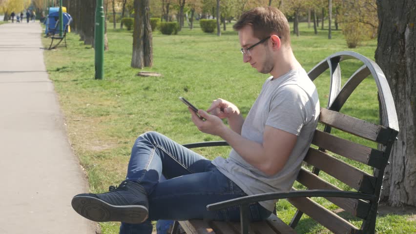 Man communicates via smartphone. Adult man sits on the bench in city park and speaks on smartphone. Man sits on the bench in public garden and uses cell phone Royalty-Free Stock Footage #1010210144