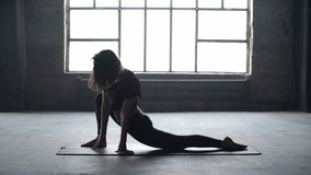 Wonderful girl with a tattoo in a dark sportswear engaged in yoga on the windows background in the loft style hall. She is standing in a half split on the mat and doing deflections, tilts and twists.