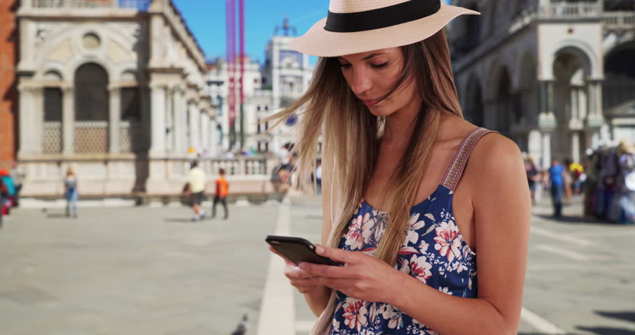 Millennial girl in romper and fedora texting on phone at St. Marks Square. Attractive girl in her 20s using smartphone to message friends while in Venice, Italy. 4k