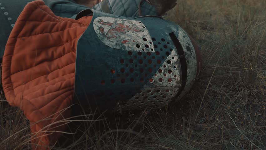 The knight in armor lies on grass with his sword and scratched color helmet