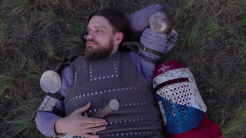 The knight in armor lies on grass with his sword and scratched color helmet