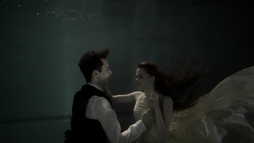 Brunette woman and young man are floating underwater. They are dressed, embracing to each other and watching tenderly