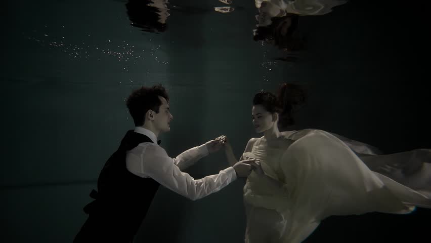 Couple of young man and woman is staying afloat under water surface in a pool. They are wearing clothes, looking to each other with love