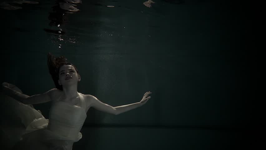 Mysterious girl with red hair is swimming in water of dark lake. She is touching surface from down and holding skirt of long dress
