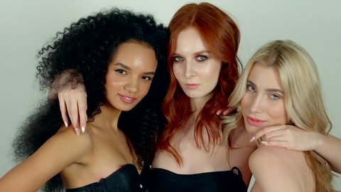 Three slim graceful women are posing for a camera and embracing. Redhead girl is hugging her friends and touching their long black and blonde hair