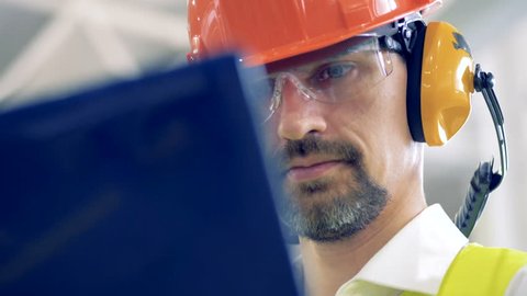 A man in hard hat works on his laptop at a warehouse, close up.