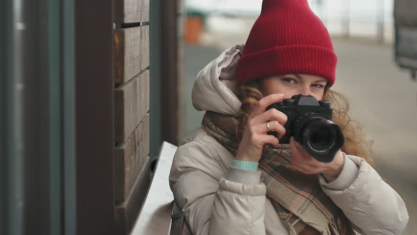 Young beautiful woman in a red hat wearing sporty warm clothes and rollers, sitting on a wooden bench and taking pictures on a vintage camera 4k