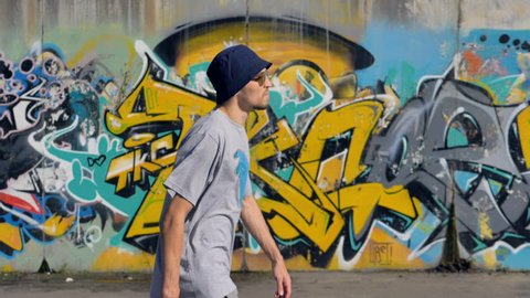 Young man is walking along the street with graffiti painting on the background and a spray in his hands