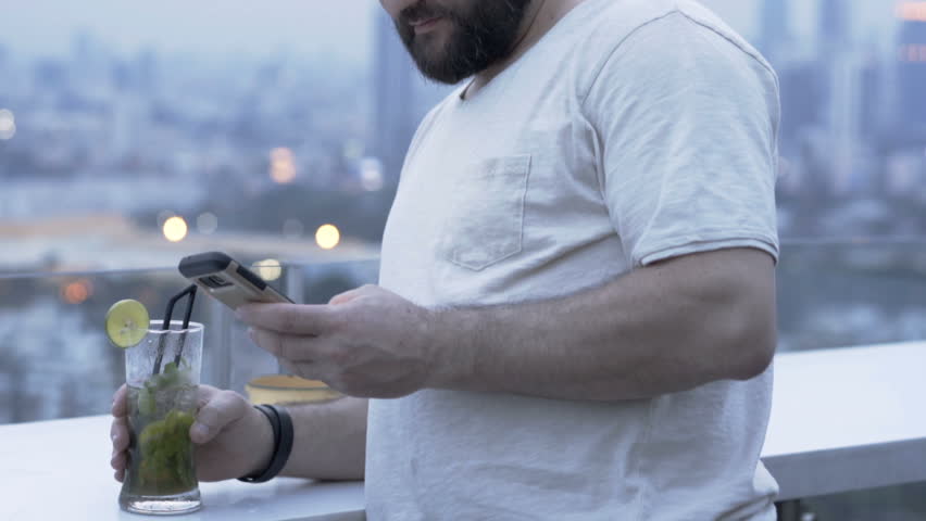 Relaxed man drinking alcoholic drink on the roof and browsing internet on smartphone
