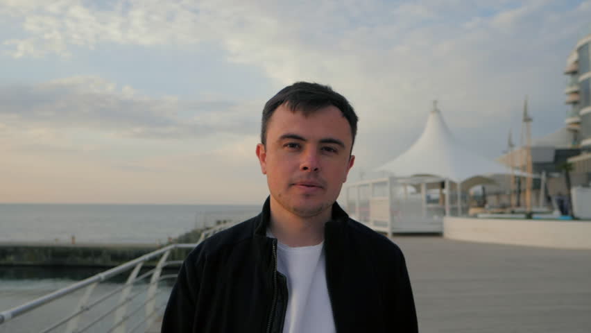 Young trendy modern man walking to camera along wooden embankment near sea or ocean. Man in fashion black bomber and white shirt. Sunset background. Slow motion