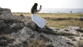 Girl takes photo near the blue sea sitting on a mountain. Woman Watching Sunset. Takes Panorama Photos On Phone. Hair in the Wind. Smartphone