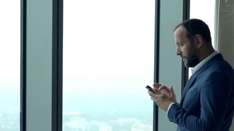Young businessman texting on smartphone standing by window in office
