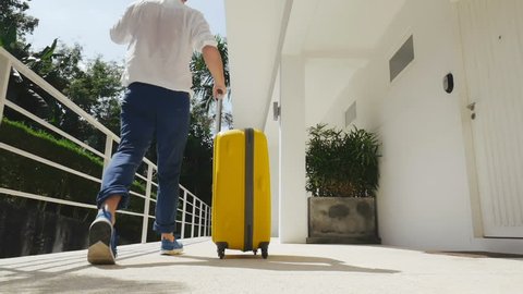 Businessman leaving modern house with a yellow suitcase. Moving camera following man talking on phone. Slow-motion.