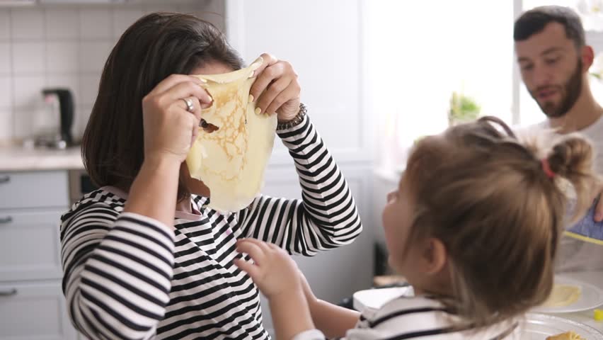Funny mother is playing with her little daughter making comical mask with pancake on her face. Family breakfast Royalty-Free Stock Footage #1010289014