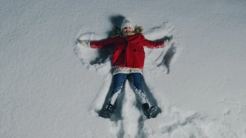 Top Down Footage of the Happy Cute Little Girl Falling into Snow, Lying and Making Snow Angel. Child Enjoying Winter Weather. Shot on RED EPIC-W 8K Helium Cinema Camera.