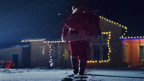 Low Angle Footage of Santa Claus with Red Bag, Walks into Front Yard of the Idyllic House Decorated with Lights and Garlands. Santa Bringing Gifts and Presents at Night.  Shot on RED EPIC-W 8K.