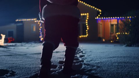 Low Angle Footage of Santa Claus with Red Bag, Walks into Front Yard of the Idyllic House Decorated with Lights and Garlands. Santa Bringing Gifts and Presents at Night. Shot on RED EPIC-W 8K Camera.