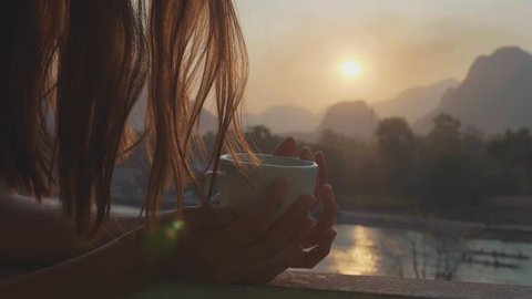 Silhouette of young woman standing on the balcony, watching the sunset and drinking hot coffee with beautiful mountain landscape on the background, slow motion.