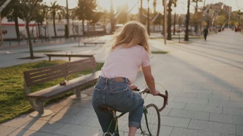 Slow motion of beautiful pretty young girl, woman with blonde hair look back at camera with happy, dreamy and romantic eyes, she rides bicycle on sunset filled park, concept forever young, summer