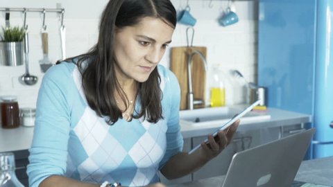 Young woman with beverage using tablet and laptop in kitchen

