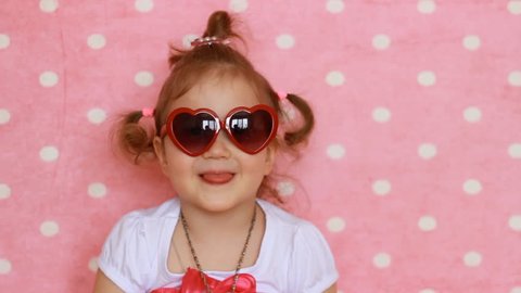 Funny child smiles, laughs and shows tongue. Pink background. Cute little girl in red sunglasses in the shape of hearts. Glamour. Fashion