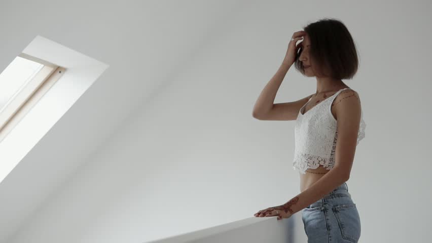 Charming woman stands in a room in scandinavian style. She is standing by the wall in a white room smiling and touching hairs. Beautiful model.