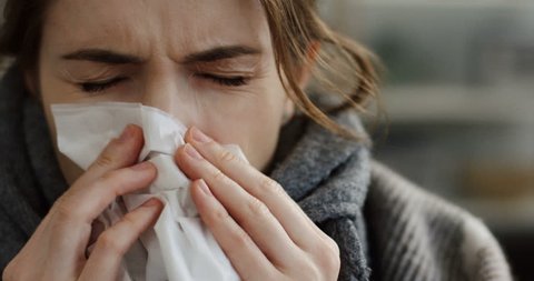 Close up of the young unhealthy caucasian woman in the warm scarf and a plaid blowing her nose in the napkin as she having a cold. At home. 2019-ncov , coronavirus , virus 