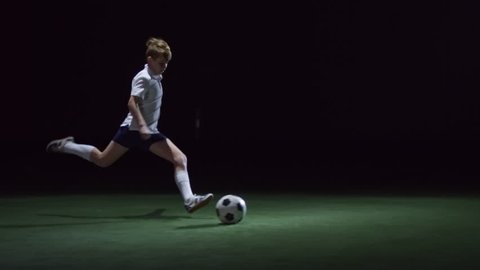 Junior soccer player running on artificial turf in indoor arena and kicking ball with power in low key lightning