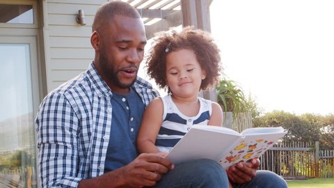 Black father and young daughter reading a book outdoors