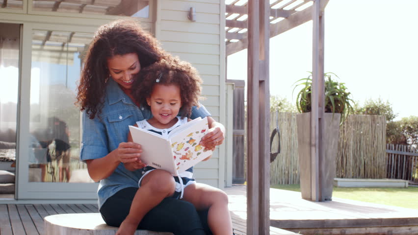 Young black girl reading book sitting on mum’s knee outdoors