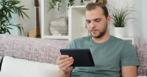 Sad Single Man Use Computer Tablet for Watching a Dramatic Movie Sitting on Sofa