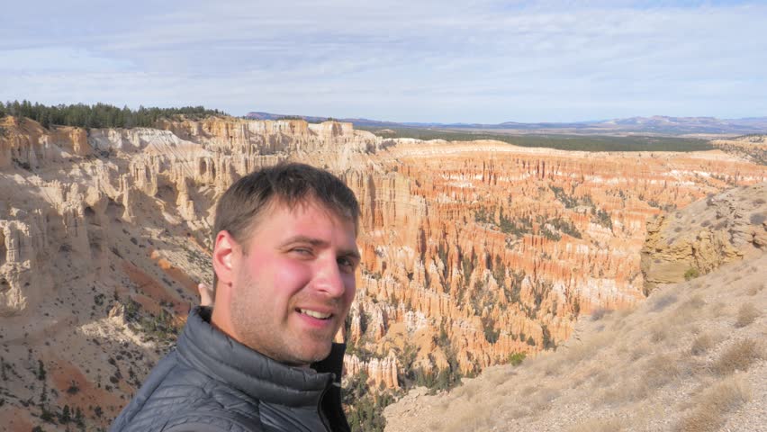 A Caucasian man with dark hair and bright white teeth smiles, while taking a selfie on the sunny Bryce Canyon rocks background. Slow motion, 4k, 3840x2160.