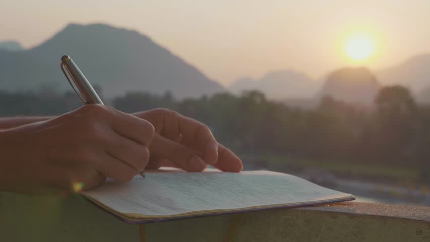 Young woman writing travel notes in diary during sunrise with beautiful sun light and mountain landscape on the background, close-up in slow motion.