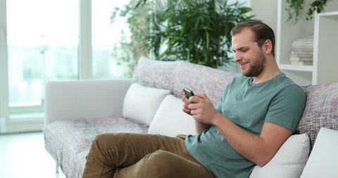 Attractive Man Play Games and Win on Mobile Phone Sitting on Sofa in Living Room