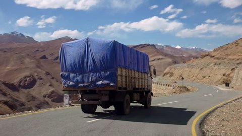 Camera follows and passes car or van driving along bent road flanked by beautiful rocky Himalaya mountains against blue cloudy sky on background. Truck ride on sunny day on Leh–Manali Highway, India.
