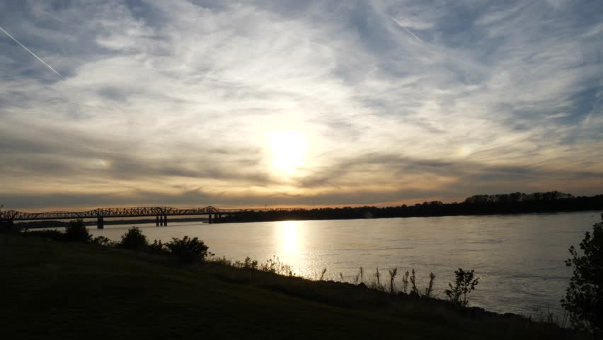 Bridge over the Mississippi River at dusk with an orange glow in the sky viewed from the bank